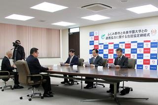 締結式にて、市長と向い合わせでスーツの男性3人が資料を持って座っている写真