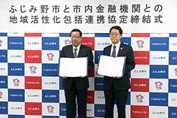 市長と眼鏡をかけて紺色のネクタイを身に着けている市内金融機関代表の方が締結書を持って横並びで立っている写真