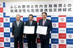 左側にグレーのネクタイを身に着けた市内金融機関の方、真ん中に市長、右側にピンクのネクタイを身に着けた市内金融機関の方が並んで立っており、市長とピンクのネクタイの方が締結書を持っている写真