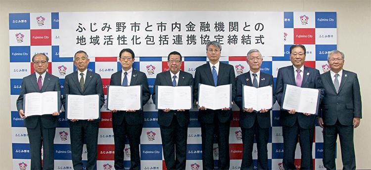 市長と市内金融機関の方々7名が横一列に並んでおり、一番右側の男性以外が締結書を持っている写真