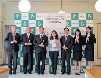 スーツを着、手にボールペンを持った男女7人が並んで記念撮影をしている写真