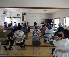 「いきいきサロン」の会場に沢山の参加者が集まって椅子に座って、前方にいる男性の話を聞いている写真
