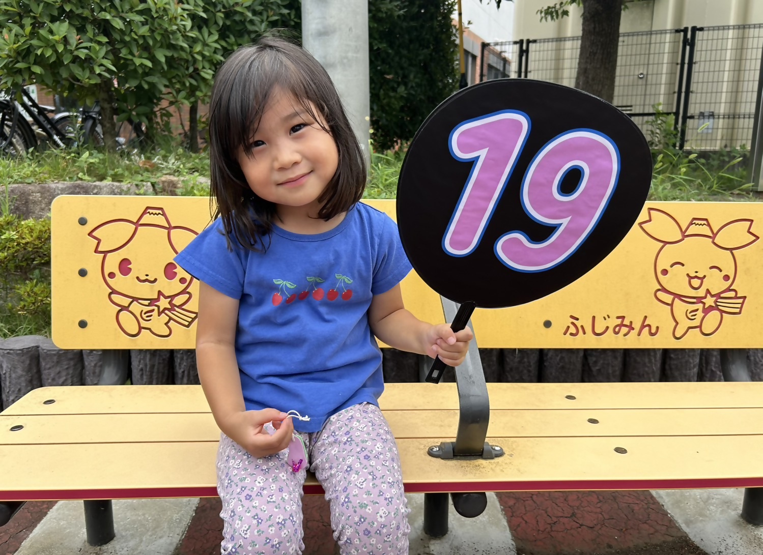 19が描いてあるうちわを持った女の子がふじみんメモリアルベンチに座ってます