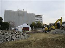 手前左側には建物を取り壊したコンクリートのがれきが積まれており、右側には砂利とショベルカーが置かれており、その奥に安全祈願祭のテントが立てられている写真