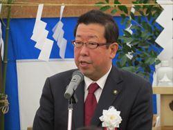 白い花の胸章を着けてマイクの前で話をしている市長の写真