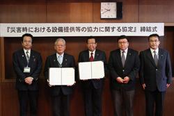 「災害時における設備提供等の協力に関する協定」締結式と書かれている横断幕の前で、市長とセンコーモータースクールの関係者の男性が協定書を持っており、関係者の方々と一緒に写っている写真1
