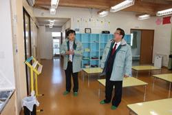 真新しい室内に机が並べられ、奥には水色の棚が置かれており、関係者の男性と市長が真新しい室内を眺めている様子の写真