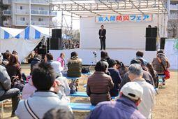 「東入間防災フェア」と書かれている横断幕のある舞台の上で市長が話をしており、ベンチに座った観客の方々が話を聞いている様子の写真