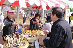 出店に団子のような食べ物と干物が竹串に刺さっている食べ物が、円形状の火鉢のようなものに沢山立てられており、市長が出店の店員の男性から品物を受け取っている様子の写真