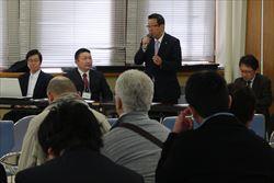 長机に座っている市長が立ち上がって、マイクを持って話をしており、来場者の方々が話を聞いている様子の写真