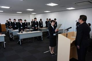 入庁式にて、新規採用職員の代表の女性が市長の前に立って書面を読み上げており、その後で新規採用職員が起立して話を聞いている写真