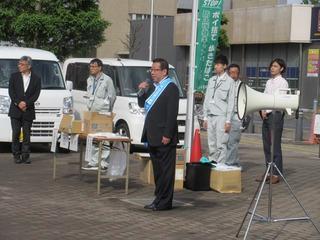 市長が水色のタスキを肩にかけて、拡声器の横でマイクを持って話をしている写真