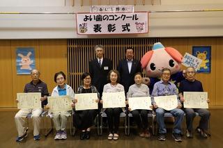 3人の男性と4人の女性が賞状を手に持って椅子に腰かけており、その後ろに市長とスーツ姿の男性とふじみんが立って写っている集合写真