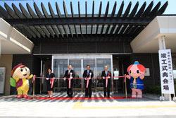 「施工式典会場」の看板が置かれている玄関前で紅白のテープリボンが貼られ、市長と関係者の方々がハサミを入れようとしており、ふじみんと黄色いふくのゆるキャラも写っている写真