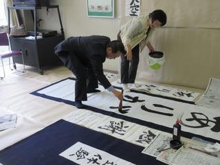 隣の女性に教えてもらいながら、大きな書道紙にありがとうと前かがみになりながら書いている市長の写真