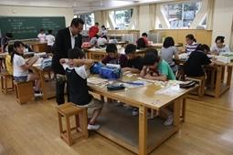 大きな机に数人ずつグループで座り1人の児童が腕を頭の後ろに組みその横に立って見ている市長を教室全体がわかるように写された写真