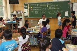 給食時間に黒板の前に男女1人ずつが立ち話をしている様子を児童や市長が見ている写真