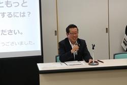 タウンミーティングにて市長がスクリーンの横の机に座りマイクで話している写真