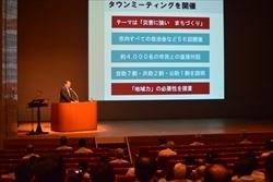 プロジェクタースクリーンの横の演台に立つ市長の写真