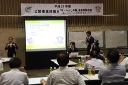スクリーンとホワイトボードの前でマイクを持った男性が会議を進めているのを聞いている参加者の写真