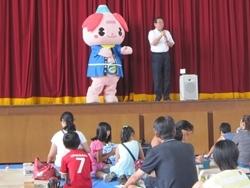 舞台で参加者の前で挨拶する市長の隣に法被姿のゆるキャラが立っている写真