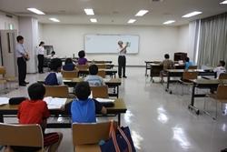 壁に立てかけられたホワイトボードの前で話しをしている講師の男性を、着席して熱心に聞いている子供たちの写真
