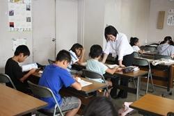 着席して学習する子供たちの席の前に立っている女性講師の写真