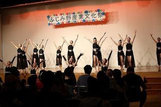 舞台上で、黒いドレスを着た女性のグループが両手を上に挙げて、ダンスを踊っている写真