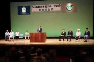 「ふじみ野市市民スポーツ大会 総合開会式」の看板が掲げられている舞台の演壇で話をしている市長と舞台上の椅子に腰かけている8名の来賓の写真
