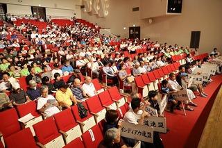 各チームの名前のプラカードを持った代表者や来場者が席に着いている会場の観客席の写真