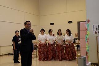 市長がマイクを持って話をしており、その後ろで、赤い花柄の長いスカートを着た女性4人が立っている写真