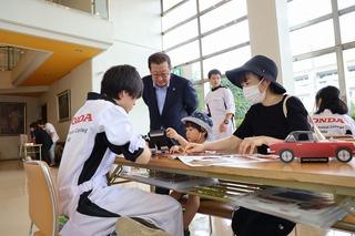 ホンダ学園の関係者の方が座っている長机に車のレプリカが置かれ、その机に男の子が車のおもちゃを持ってお母さんと座っており、男の子の様子を市長が後ろから見ている写真