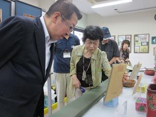 作品が展示されており、写真たてのような作品を黄緑色の服を着た女性が手に取って市長に説明をしようとしている写真