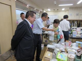 バザー会場で机に沢山のバザーの品物が並んでおり、買い物に着た女性が品物を選んでおり、隣にいるワイシャツを着た男性が女性に話かけている様子を市長が笑顔で見ている写真