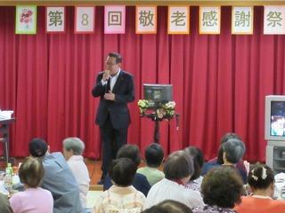 「第8回敬老感謝祭」の横断幕の貼られている舞台上で市長がカラオケを歌っており、出席者が座って聞いている写真