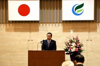 日の丸の国旗と市旗が掲げられており、その前の演台で市長が話をしている写真