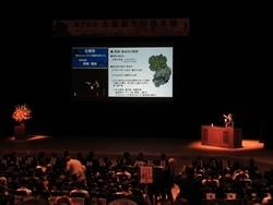 スクリーンが映し出されたステージの演題で発表している市長の写真