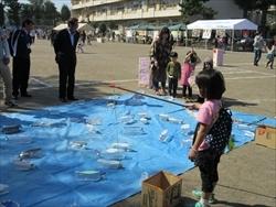 グラウンドに設置されたブルーシートの上で魚釣りをしている女の子を親子連れが見ている写真