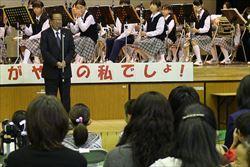 環境フェアの舞台で市長が来場者に話をしており、市長の後ろには吹奏楽部の学生が写っている写真