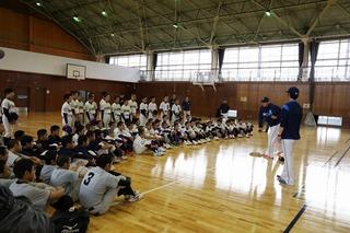 オープニング式典で野球のユニフォームを着た人達が座りながら、真ん中で話をしている人を見ている全体写真