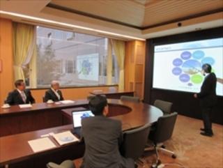 株式会社KDDI研究所にて、市長と塩川 修埼玉県副知事が、前方の大型スクリーンの映像を見ながら説明を受けている写真