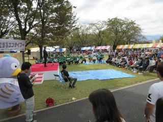 舞台前の芝生の上にブルーシートが敷かれて、その周りに並んで話を聞いている参加者の方々の全体を写した写真