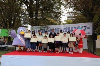 舞台上でマスコットキャラクターと一緒に子供達が市長と集合写真