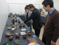 机の上に沢山の焼き物が展示されており、市長が急須を指差して、隣に写っている女性と話をしている写真