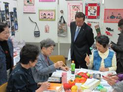 手作り品の展示コーナーの前の机に、年配の女性が集まって座っており、机の上に広げた白い紙に筆を持って絵を描いており、その様子を後ろから市長が見ている写真