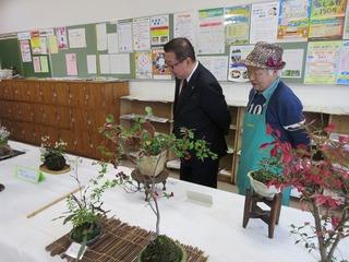 帽子に緑のエプロン姿の男性と一緒に、机に並べられた作品を見学している市長の写真