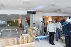 新しいく出来上がっているフロアの写真で、手前には沢山のダンボールに入った荷物が積まれており、右側には作業を行っている工事関係者と、視察を行っている市長が写っている写真