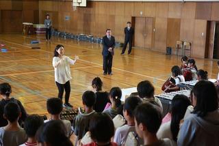 体育館で音楽会の練習をしている生徒達と指揮者の先生の後ろで見ている市長の写真