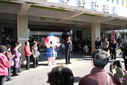 市役所の入り口前に集まる大勢の住民の前で話しをしている市長とその横でゆるキャラが手を振っている写真