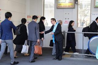 駅のホームに入って行く市民の方々に、青いタスキを肩にかけている市長がキャンペーンのチラシを手渡ししている写真